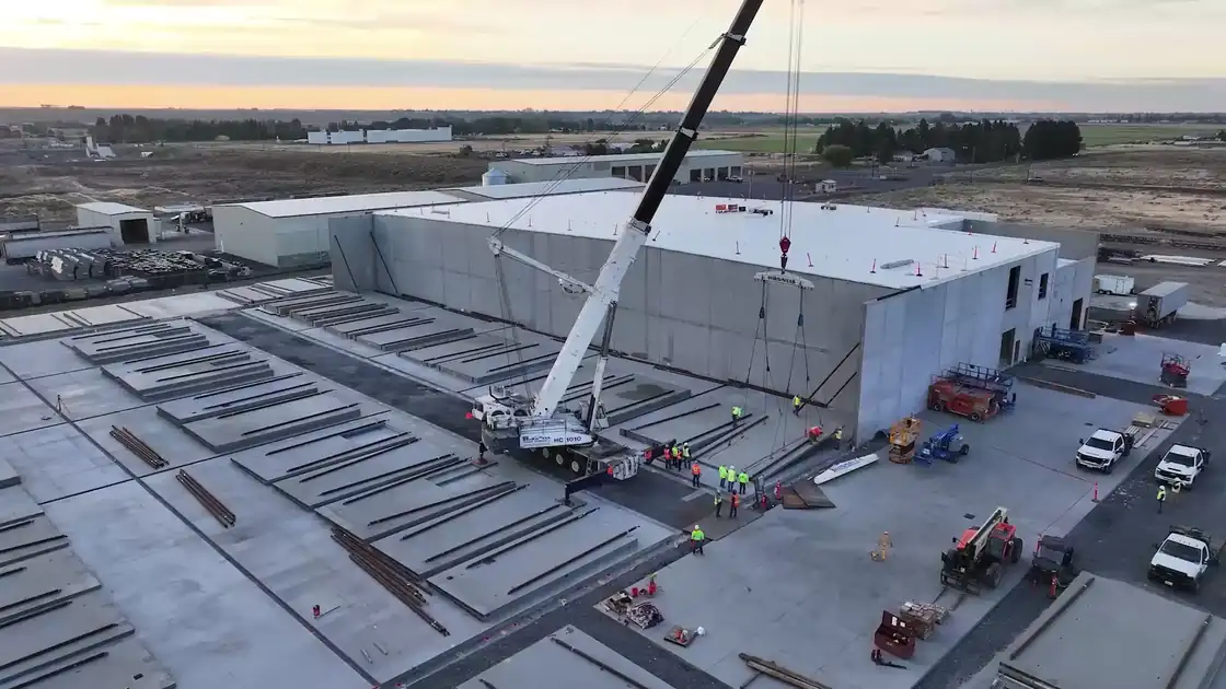 A crane lifting a tilt-up concrete wall panel into place at sunset, seen from above